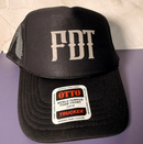 A black trucker hat with 'FDT' embroidered in white on the front, resting on a purple surface against a light beige textured background. A white Otto Trucker sticker is attached to the brim.