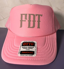 A pink trucker hat with 'FDT' embroidered in white on the front, resting on a purple surface against a light beige tiled background.